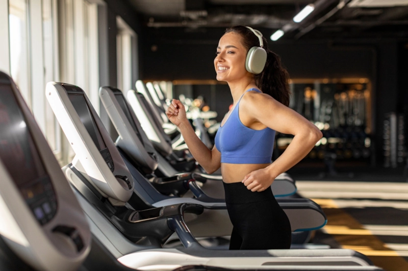 A smiling woman in headphones running on a gym treadmill