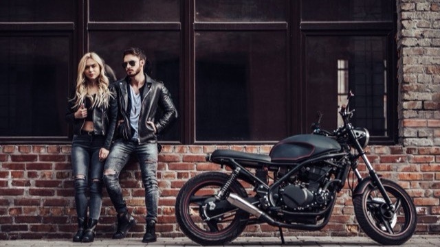 A couple in leather jackets leaning against a cafe racer motorcycle outside a brick-fronted cafe