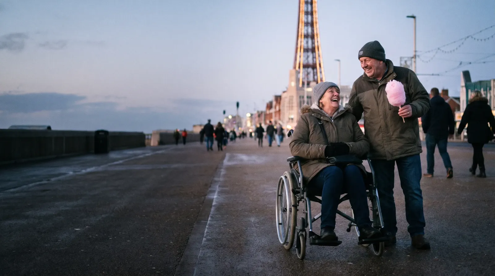 A couple on Blackpool promenade at dusk with the Tower behind; the woman is a wheelchair user and the man holds candyfloss