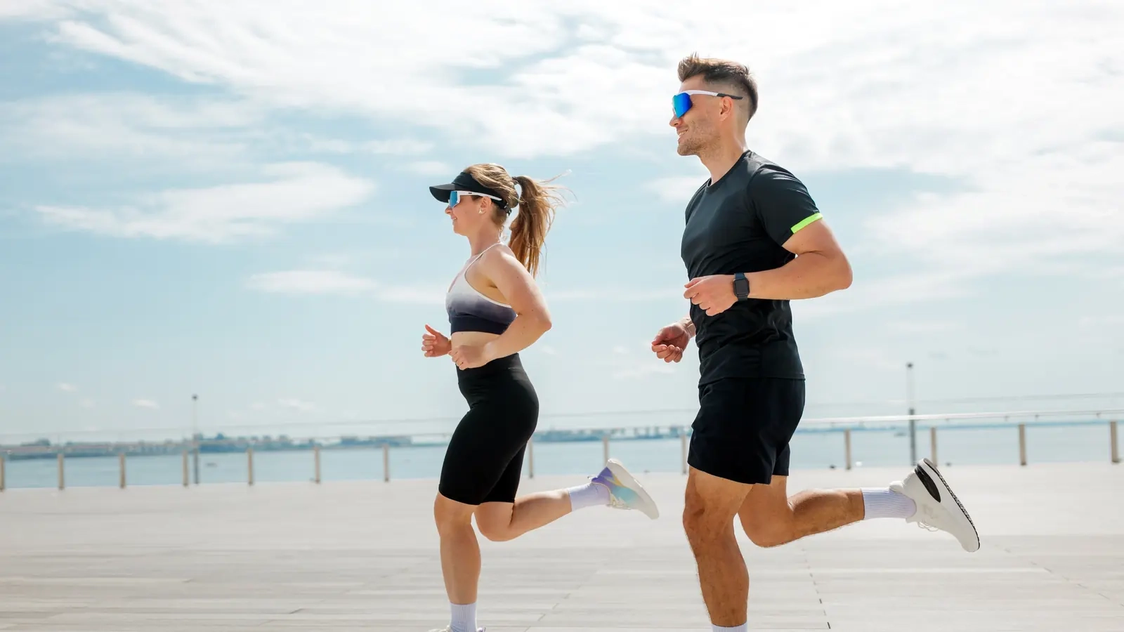 A man and woman running side by side along a sunny seaside promenade