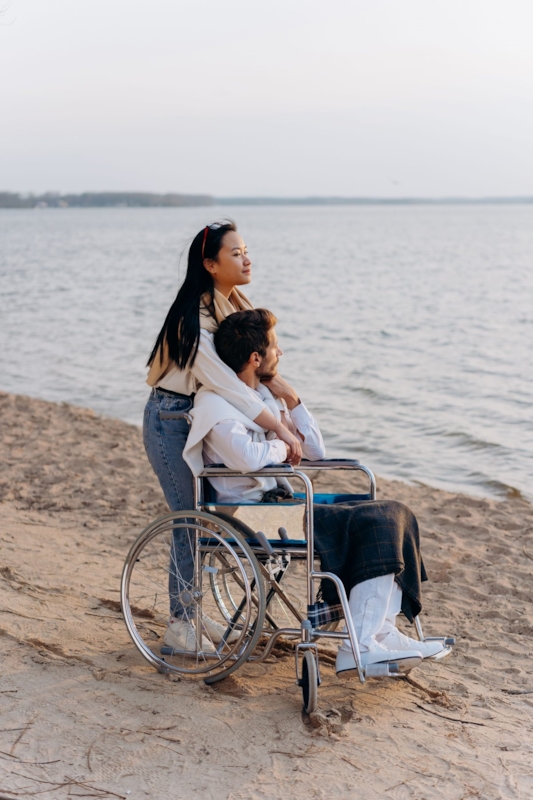 A woman standing behind a man in a wheelchair, both looking out at the water on a beach