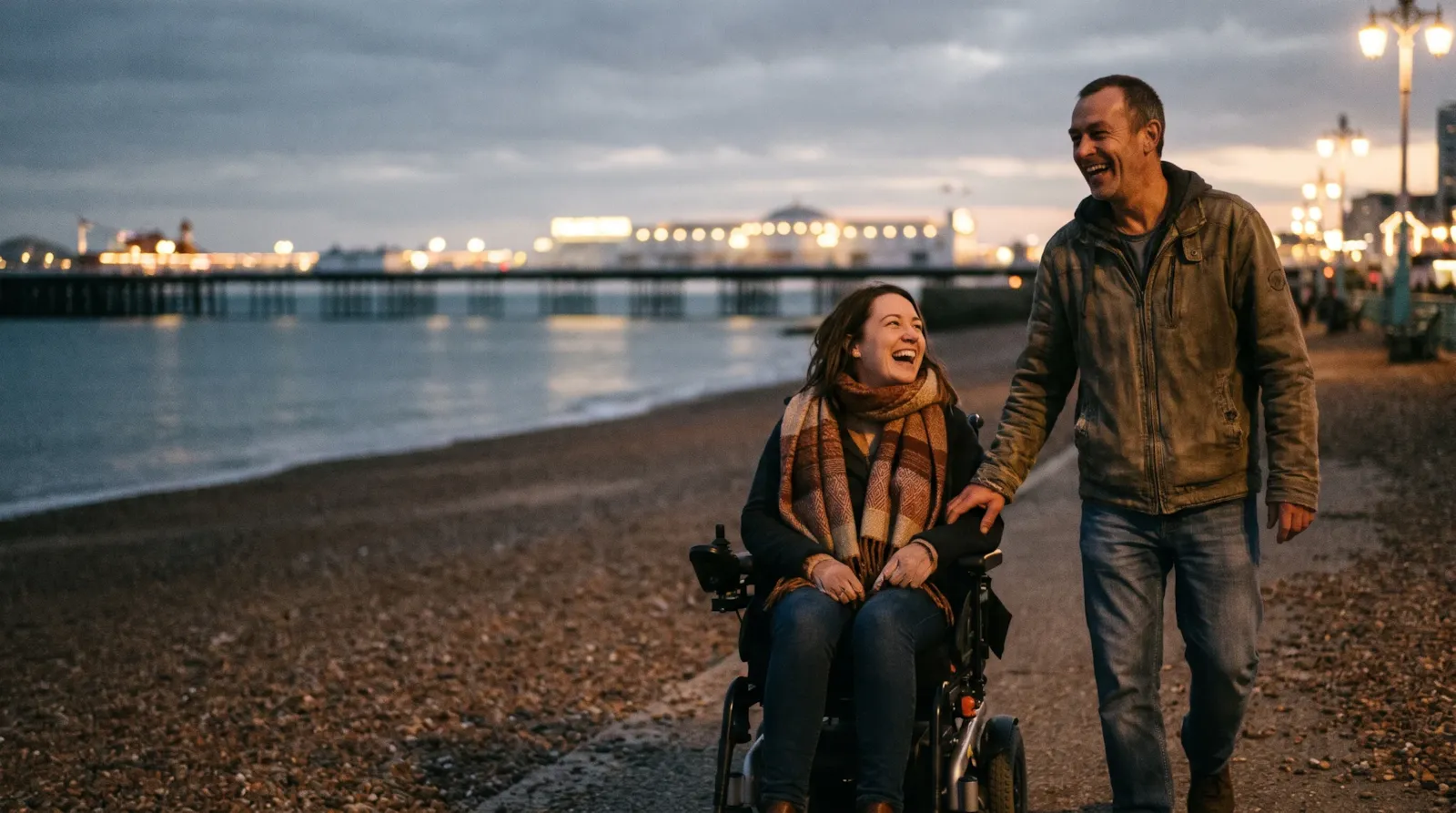 A couple laughing on Brighton seafront at dusk, the Palace Pier lit up behind them; the woman uses a powered wheelchair