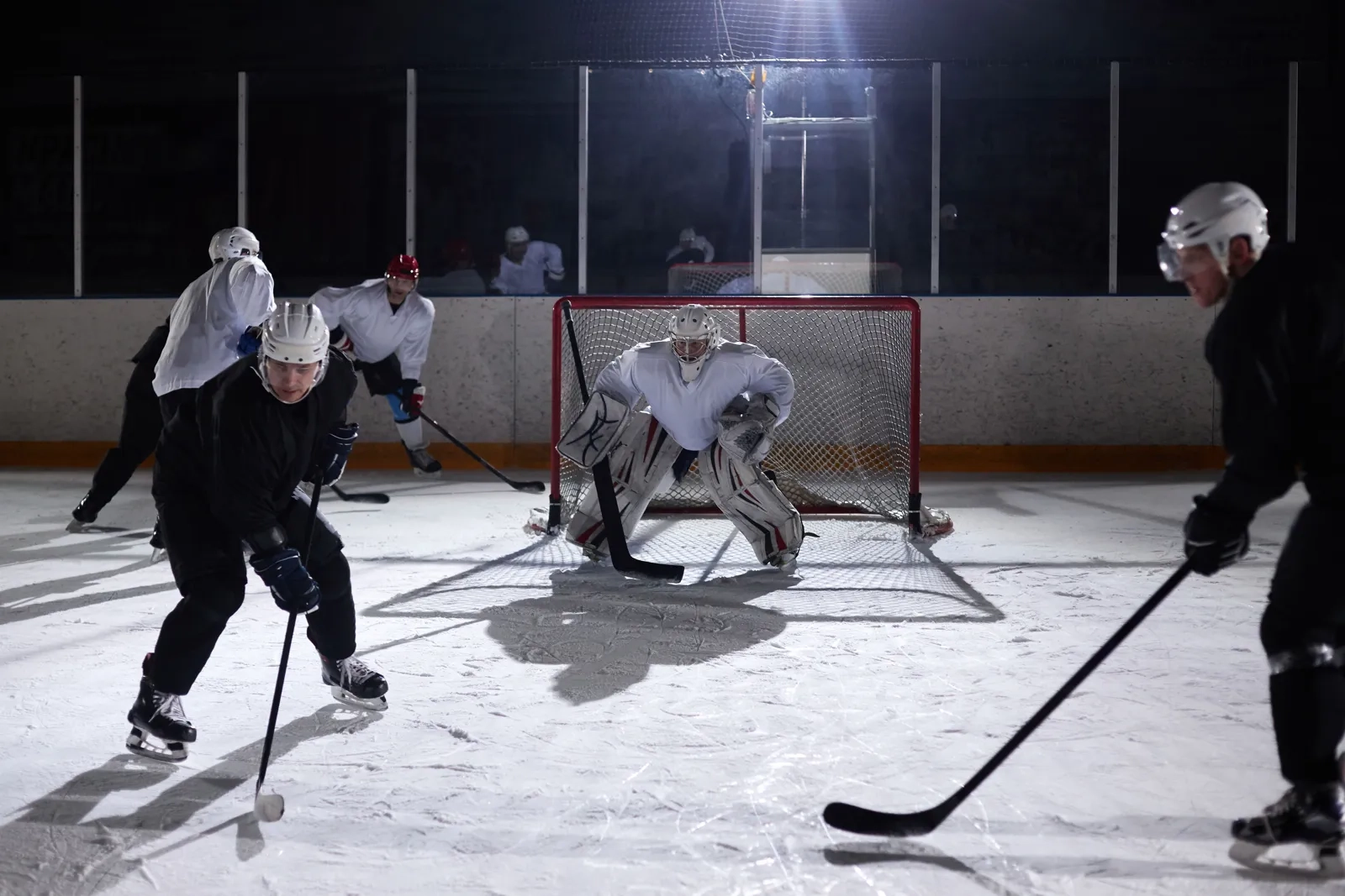 Players in dark jerseys and a goalie in front of the net during a night game at an indoor rink