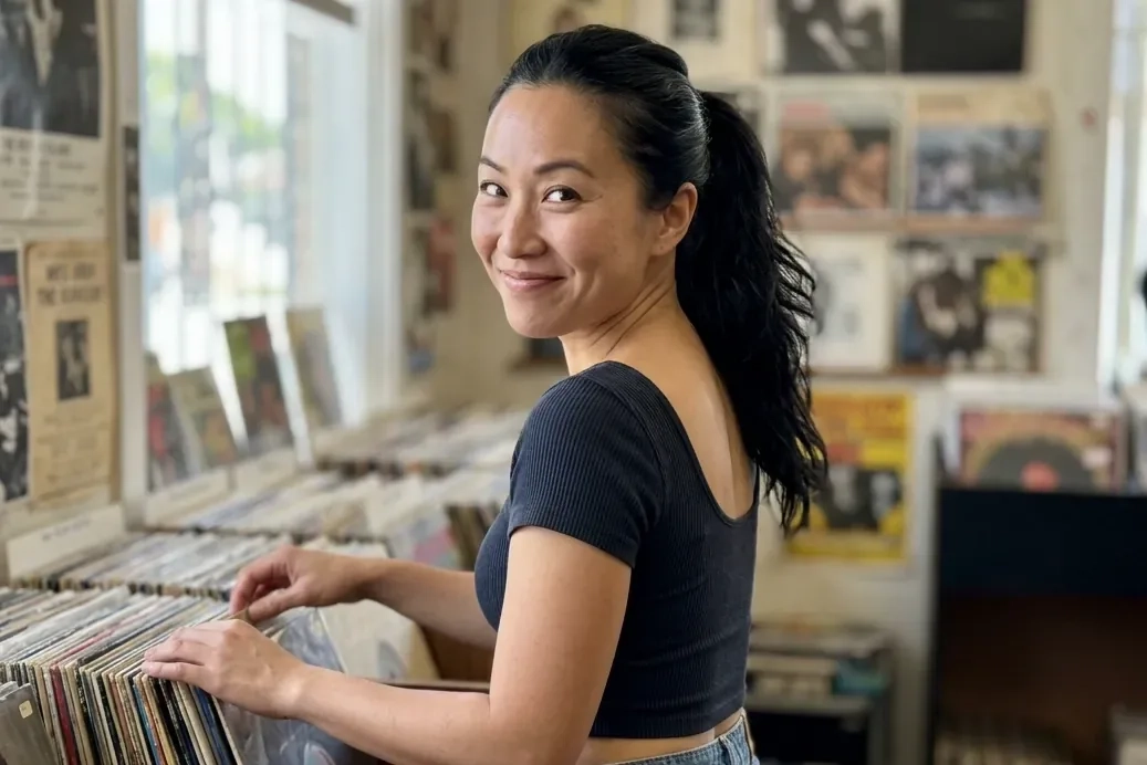 A woman with a sleek black ponytail and a fitted crop top flips through vinyl records in a record shop and smiles back over her shoulder (AI-generated image)