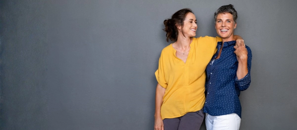 Two women stand close together against a grey wall, smiling