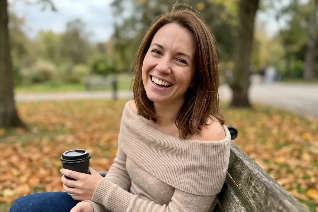 A laughing woman with chestnut hair and an off-the-shoulder sweater sits on a park bench in autumn leaves with a coffee and her phone (AI-generated image)