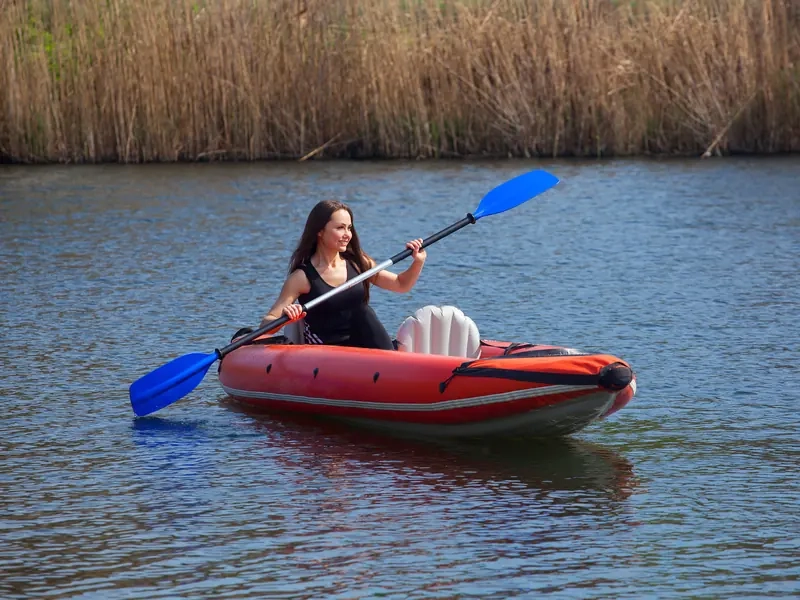 A smiling woman paddling a red inflatable kayak past tall reeds on a calm river