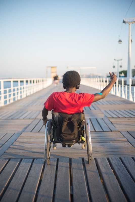 A man in a wheelchair raising his hand while looking out over the water from a boardwalk pier