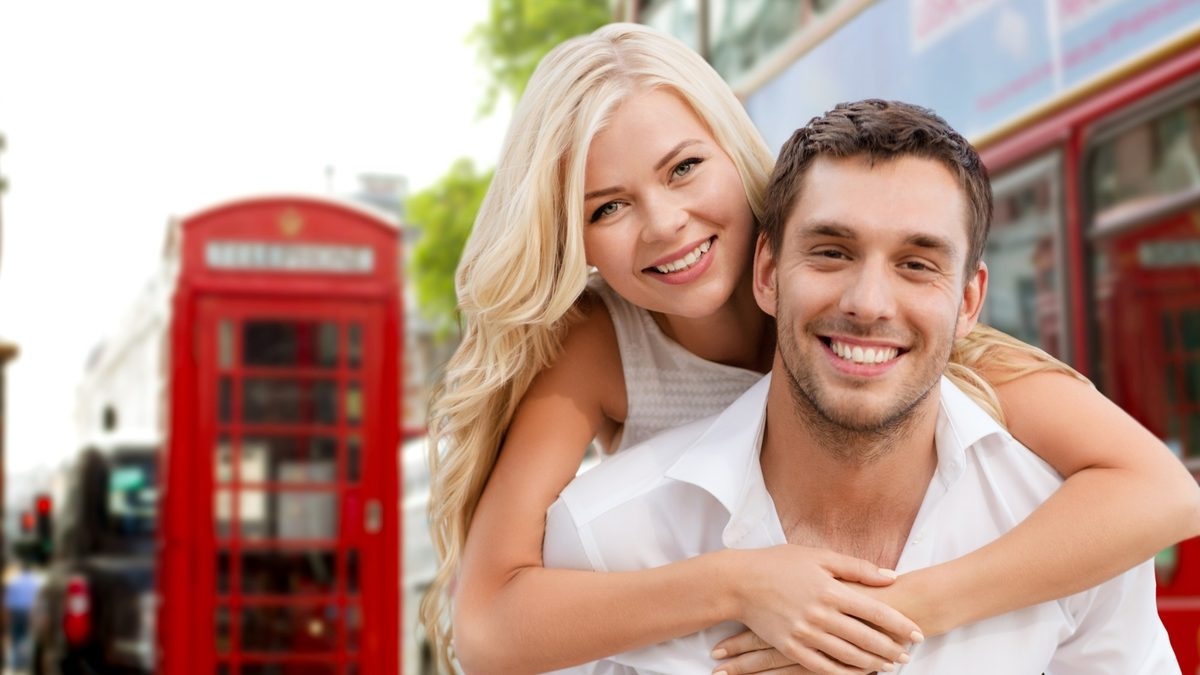 A couple smiling together in front of a red London phone box and double-decker bus