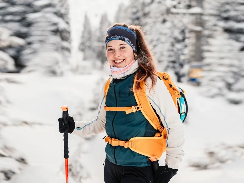 A smiling woman with a backpack and poles hiking through a snowy forest