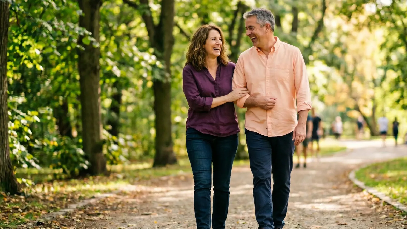 A couple walking arm in arm and laughing together along a sunny, tree-lined park path